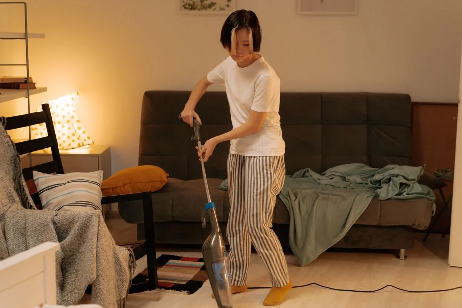 Two women in work clothes tidying a modern living room with a vacuum and mop.